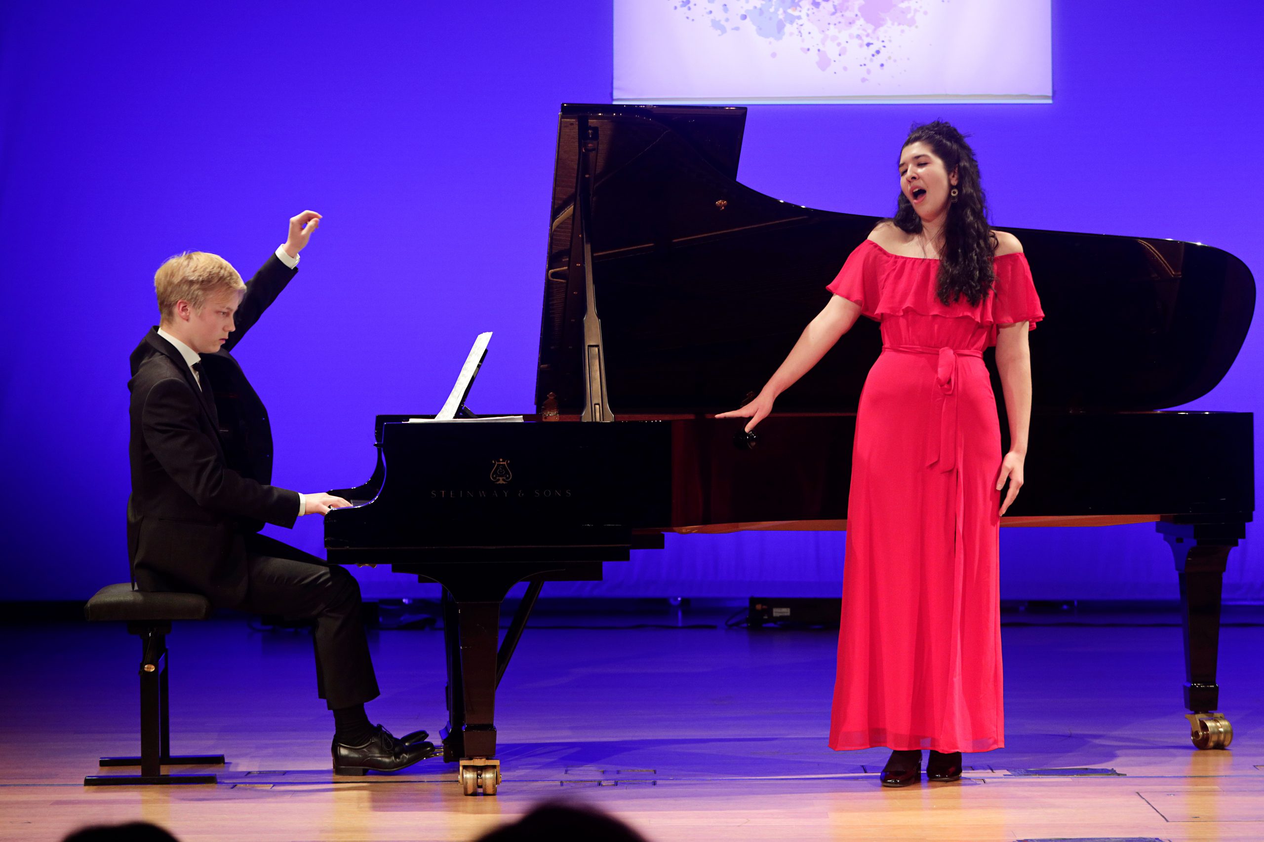 Vasemmalla mustaan pukuun pukeutunut pianisti säestää Steinway & Sons flyygelillä, oikeammalla olevaa solistia punaisessa mekossa. Kuva otettu yleisön perspektiivistä suhteellisen läheltä.